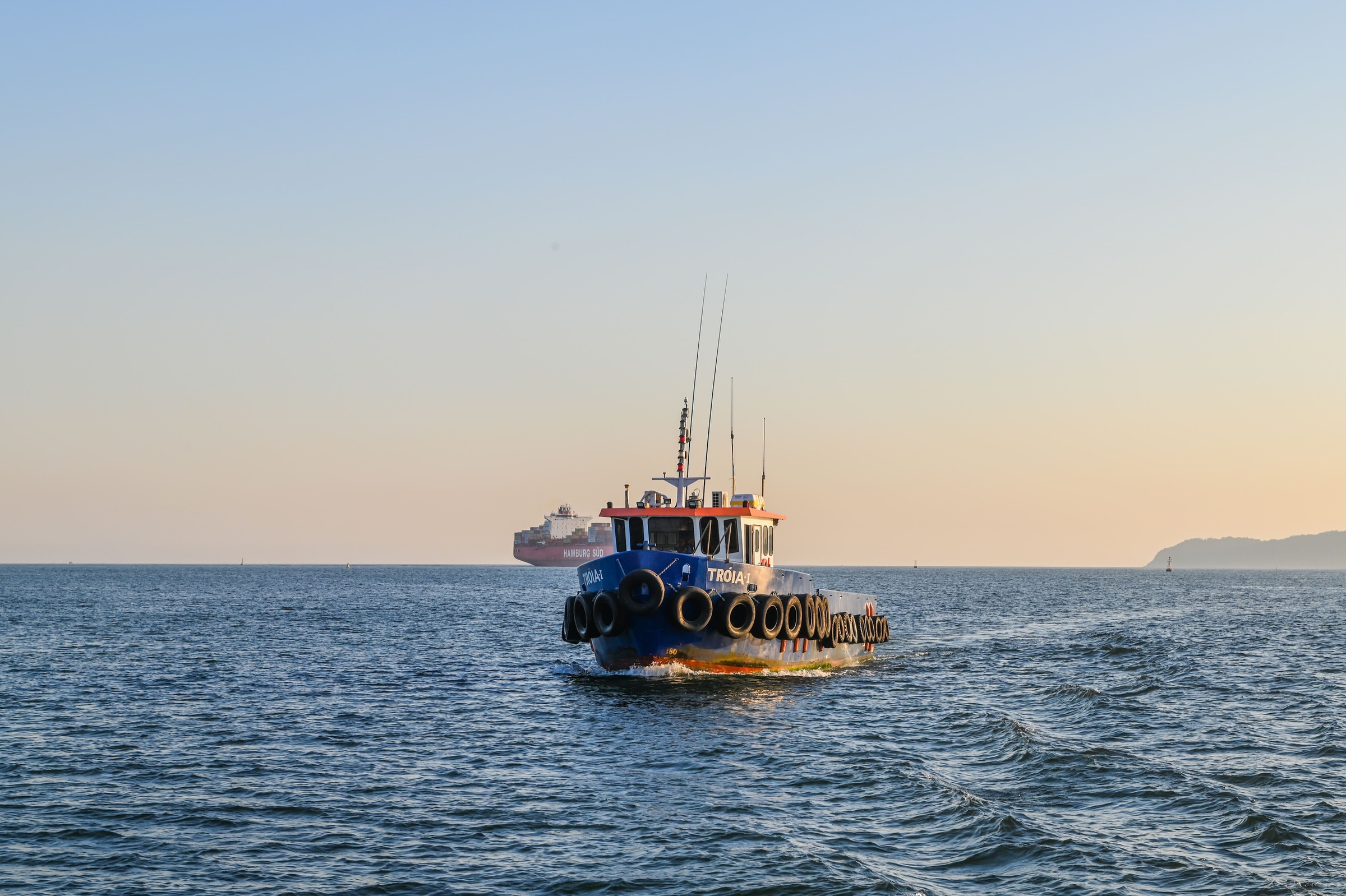Embarcação Tróia I navegando ao entardecer rumo a um navio fundeado